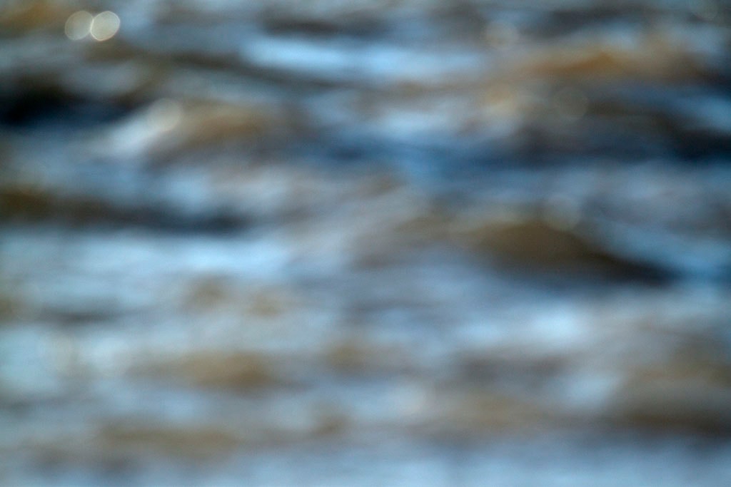 Depoe Bay, OR 2017 — blurred coastal water abstract, cool blue-grey with warm tan wave forms and single bokeh highlight upper left