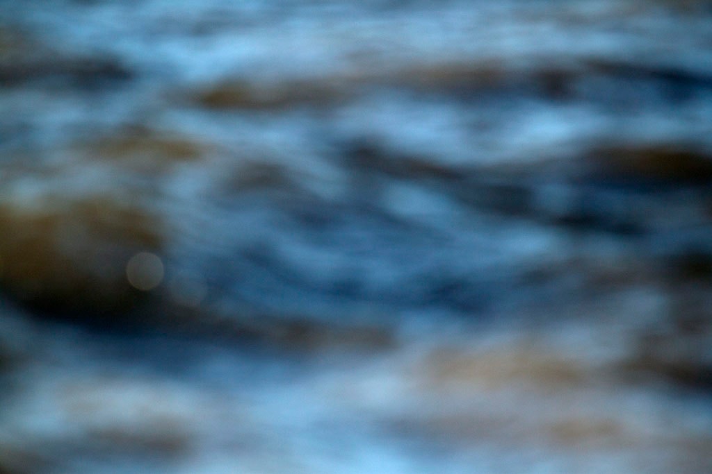 Depoe Bay, OR 2017 — blurred coastal water abstract, cool blue-grey with warm tan wave forms and single bokeh highlight lower left